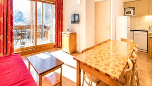 Een kamer met een houten tafel, stoelen, een televisie, een keukenhoek met apparatuur, en een raam met gordijnen.