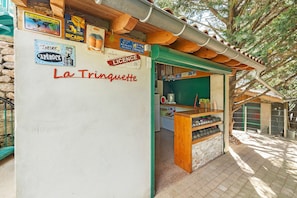 A rustic outdoor café with a wooden counter, a green awning, and various signs including 'La Trinquette.'