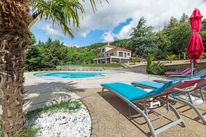A pool area with lounge chairs, a red umbrella, and a house in the background.