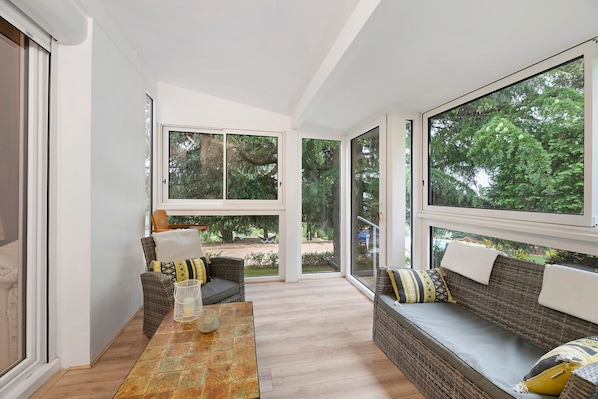 A sunlit room with a sofa, a table, and large windows offering a view of greenery.