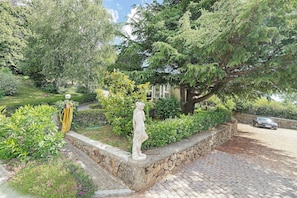 A garden with a statue, stone wall, and a paved area with a car in the background. - Chalet "Au Domaine De Cabu" with Mountain View, Shared Pool and Wi-Fi (Saint-Victor)