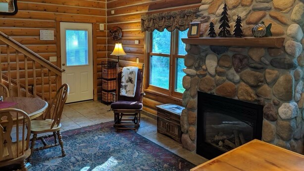 A log cabin interior with a stone fireplace, wooden walls, a dining area with a table and chairs, and a window with a view of trees. - Cozy lake cabin in Crosslake with a fireplace, perfect for relaxing getaways. (Crosslake)
