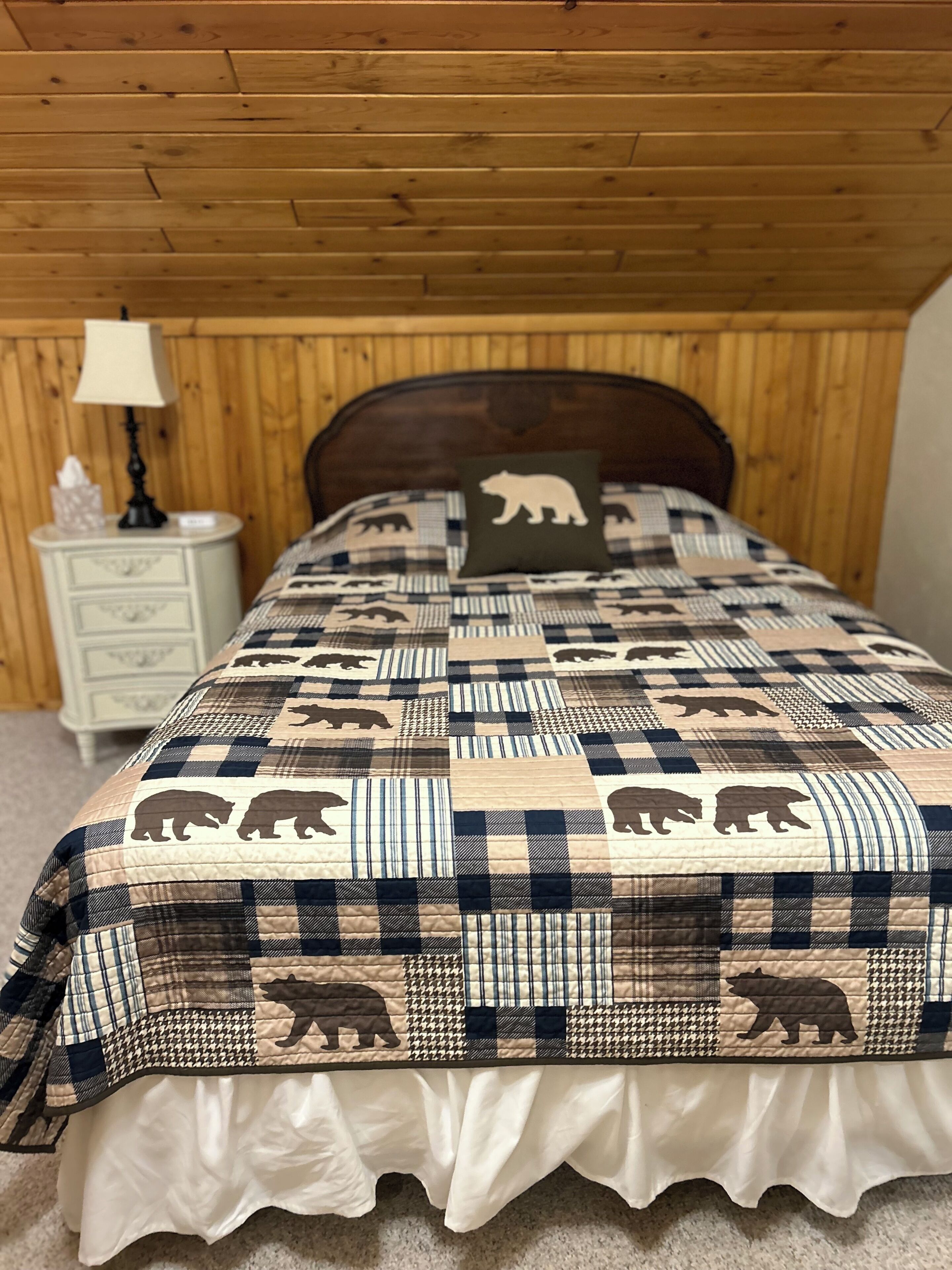 A bedroom with a wooden ceiling, a bed with a quilt featuring bear patterns, a bedside table with a lamp, and a nightstand with drawers.