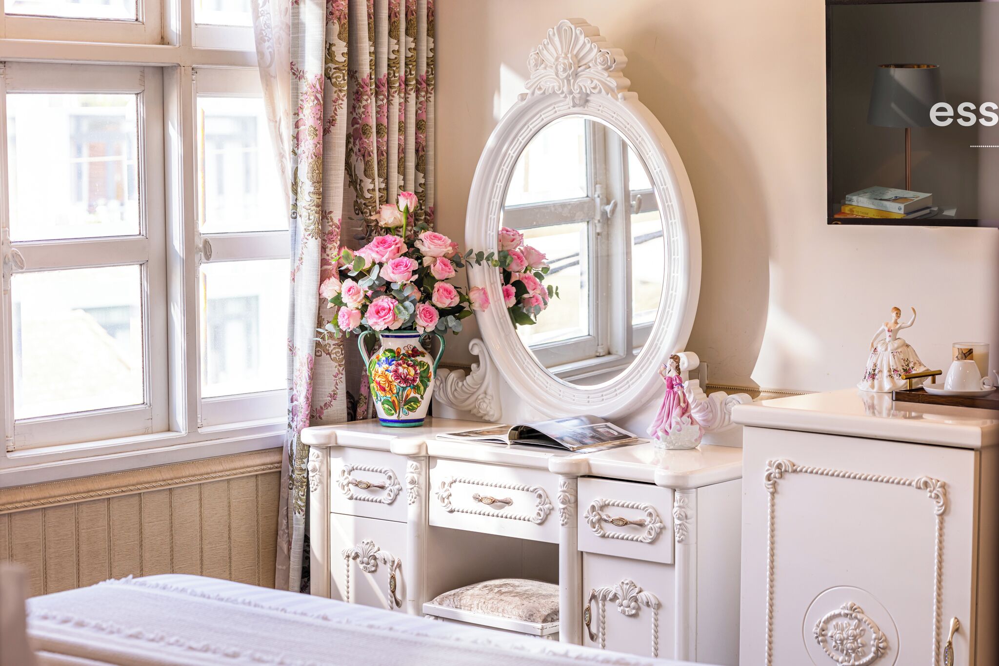 A room with a white vanity, a mirror, a vase of pink flowers, and a bed with a white headboard.