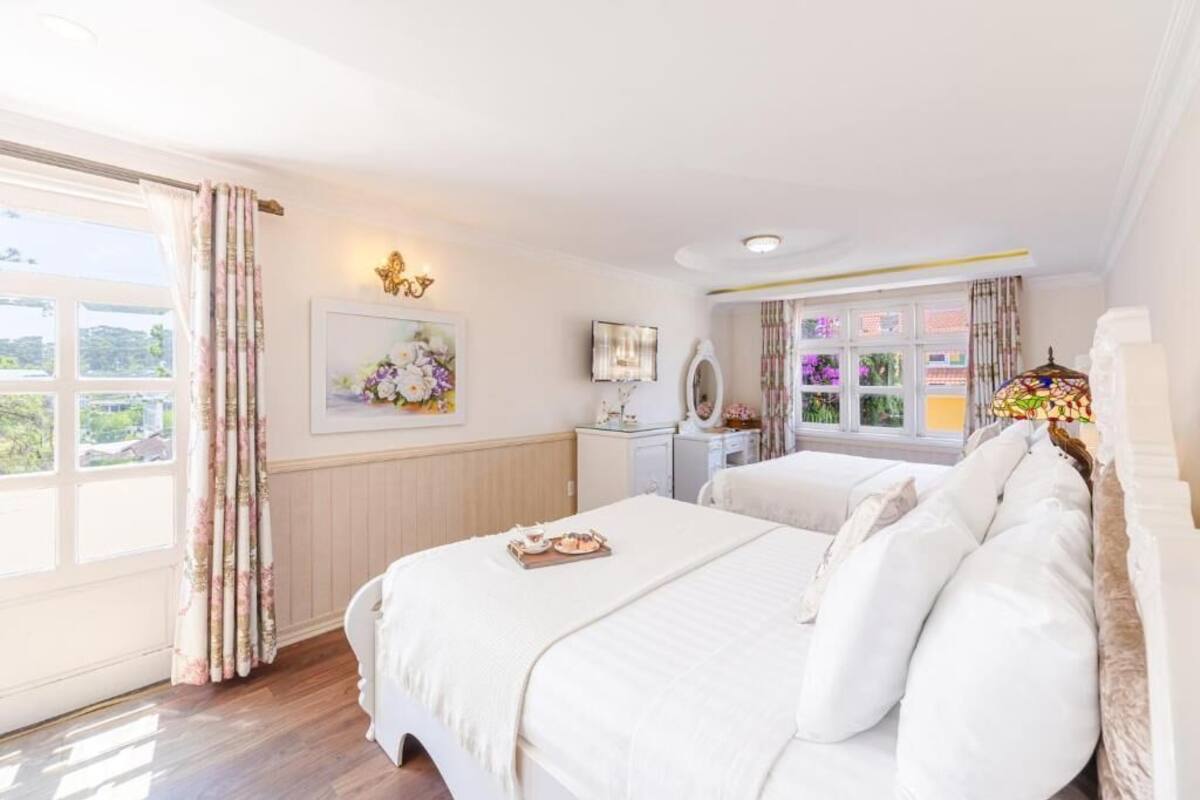A bedroom with two beds, a window with curtains, a bedside table, and a wall-mounted lamp.
