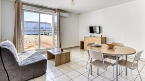 Un soggiorno moderno con un divano, un tavolo da pranzo con sedie, una TV su un mobile e un balcone con vista sugli edifici.