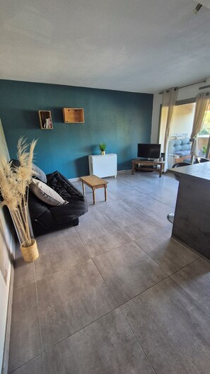A living room with a dark sofa, a small wooden table, a TV, and a window with curtains. - Apartment 2 rooms 45m2 quartier Saint Jean ANTIBES (Antibes)