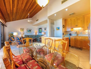 A spacious living area with a dining table, chairs, a sofa, and a kitchen area with wooden cabinets. - Makena Surf B305 Beachfront Resort (Wailea)