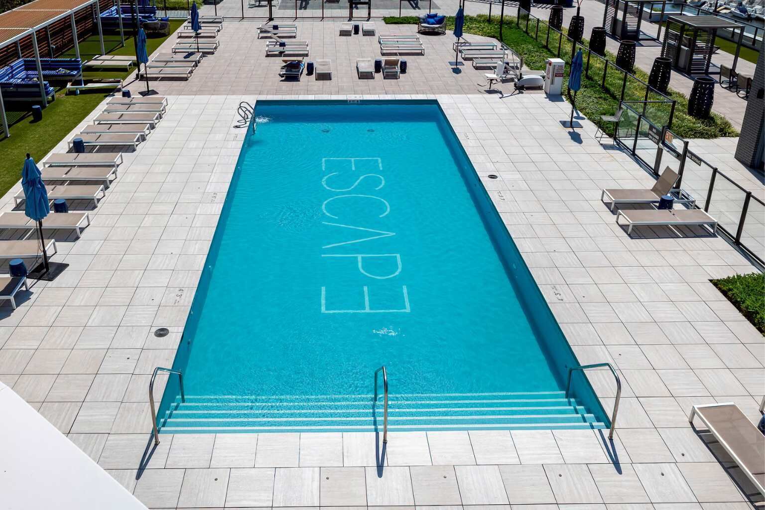 Une piscine rectangulaire sur laquelle on peut lire le mot «  ÉVASION » à la surface de l’eau, entourée de chaises longues et de parasols.