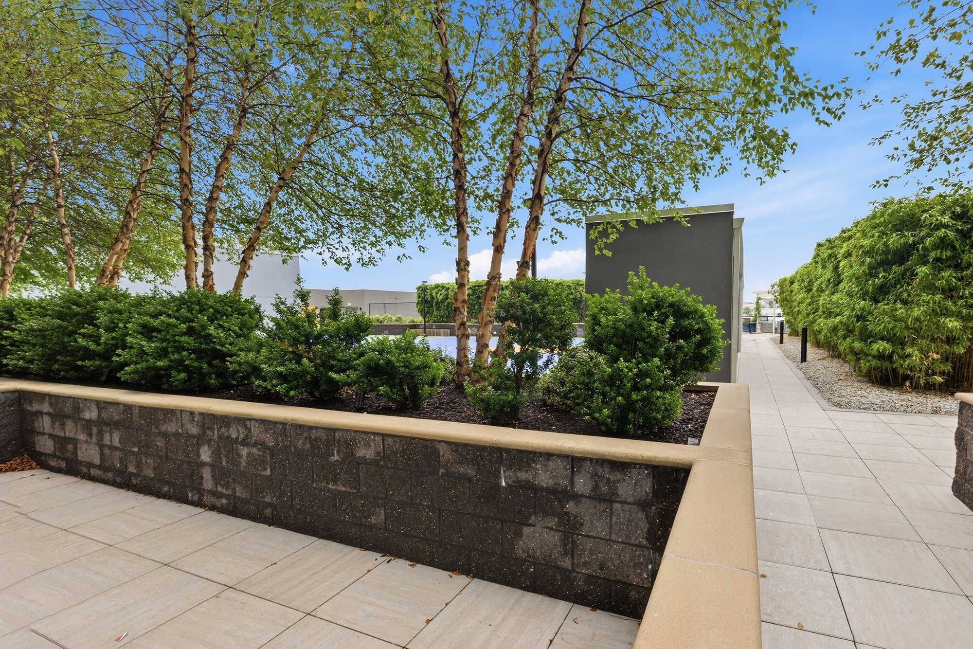 A paved walkway with a stone wall and greenery, leading to a modern building with a flat roof.