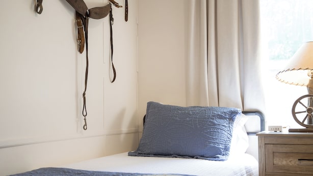 A bedroom with a bed, a bedside table with a lamp, a window with curtains, and a wall-mounted coat rack with items.