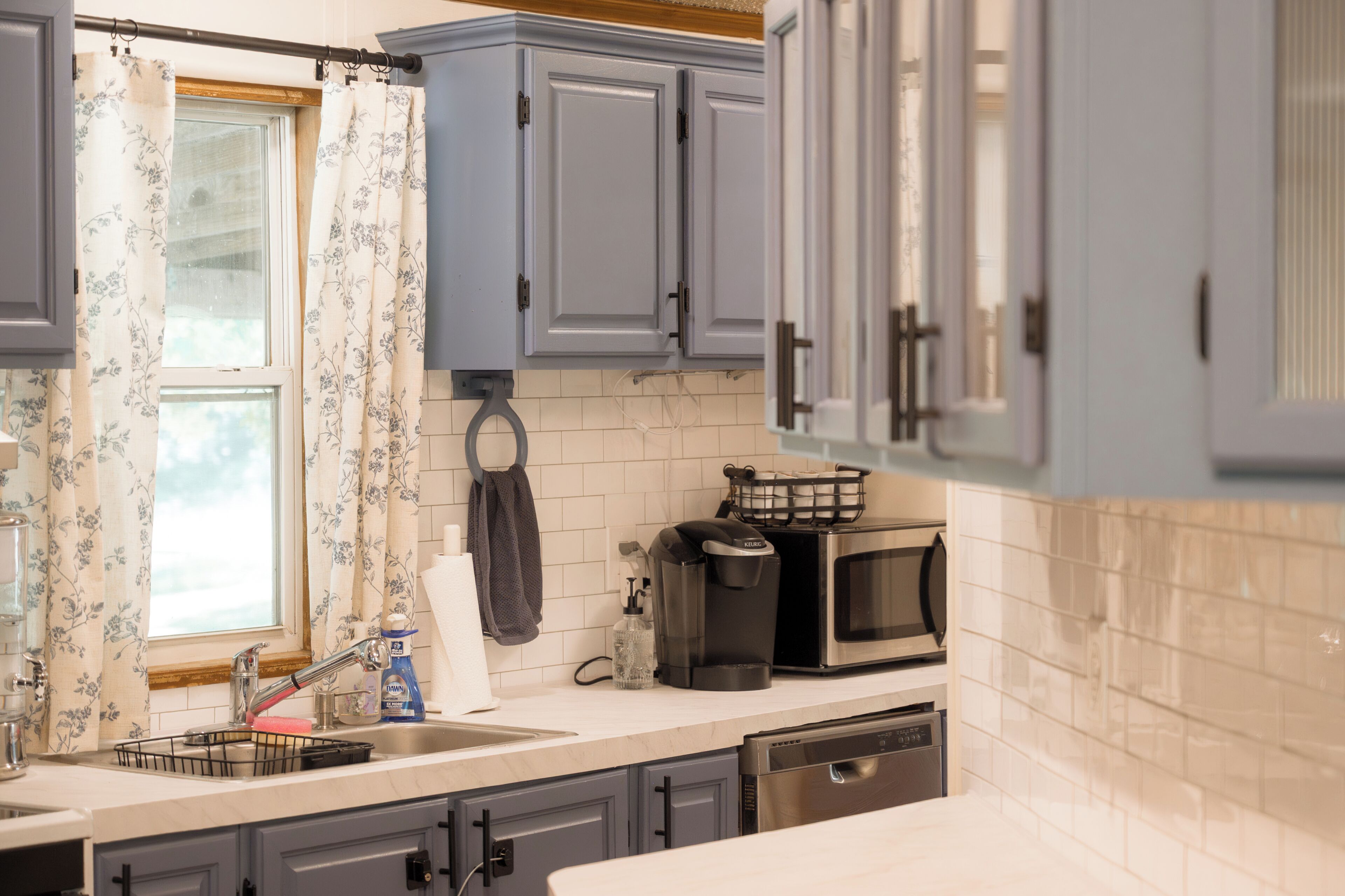 A kitchen with grey cabinets, a stainless steel sink, a coffee maker, and a dish rack.