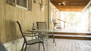 A wooden porch with a glass table and metal chairs, a bull skull mounted on the wall, and a wooden railing overlooking a garden.