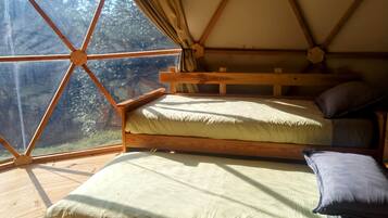 Una cama de madera con dos colchones y almohadas, un ventanal con vistas a árboles y suelo de madera.