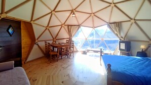 Una stanza a cupola geodetica con pavimento in legno, un tavolo con sedie, un letto con coperta blu e un televisore.