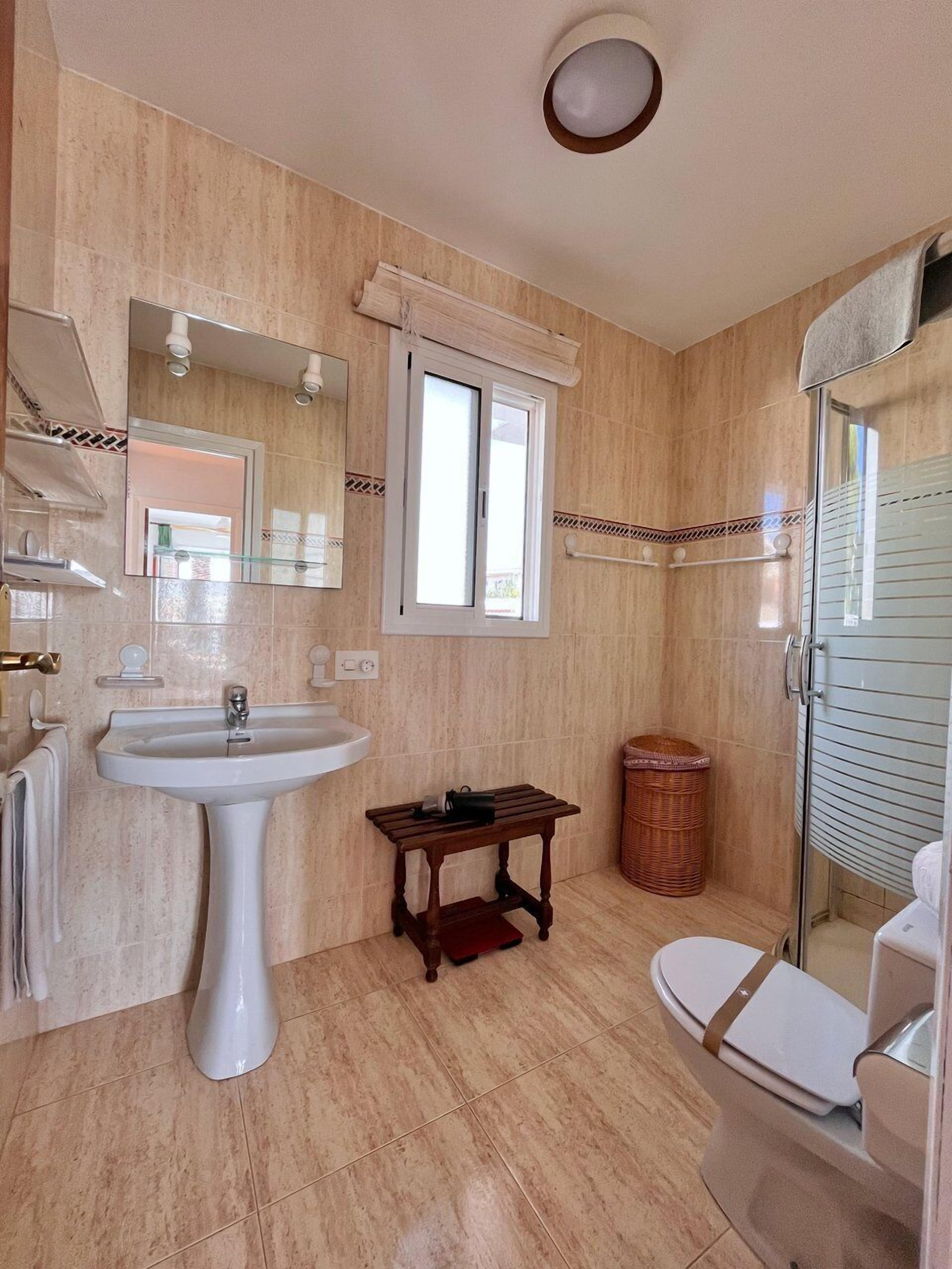 Ein Badezimmer mit einem Waschbecken auf Säule, einem Spiegel, einem Fenster, einem kleinen Tisch, einem Korbwäsche und einer Toilette.