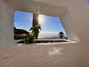 A beach view through a window, including a palm tree and a rocky shoreline. - Beachfront Getaway with Fantastic Views (L'Ampolla)