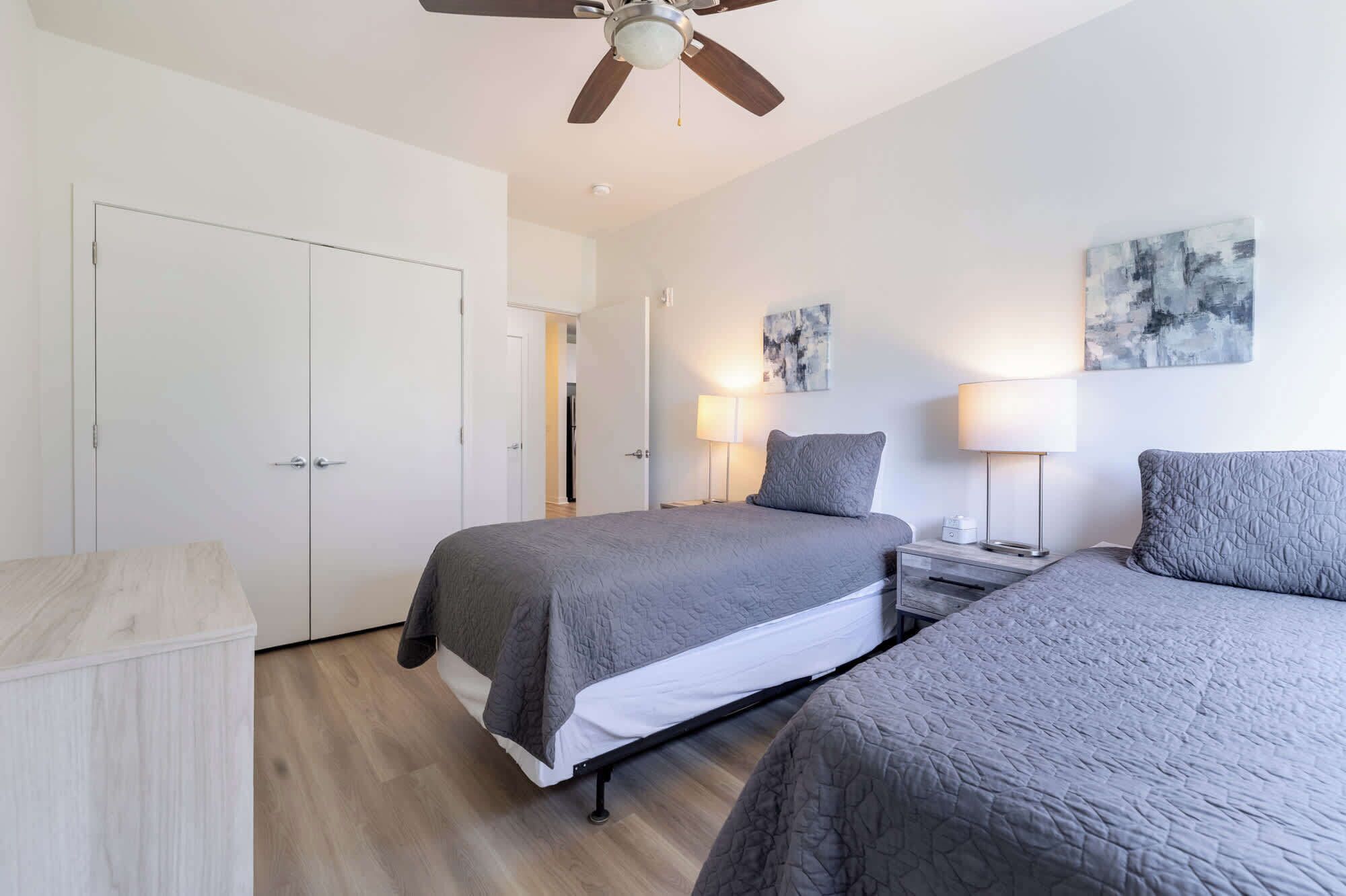A bedroom with two beds, a ceiling fan, bedside lamps, and abstract wall art.