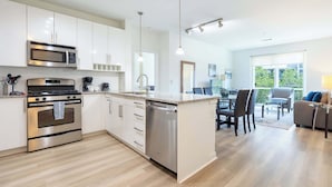 A modern kitchen with stainless steel appliances, a dining area with a glass table, and a living space with a sofa and a view of greenery.