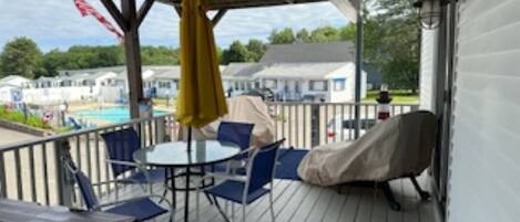 A covered outdoor area with a table and chairs, a covered grill, and a view of a swimming pool and houses in the background.