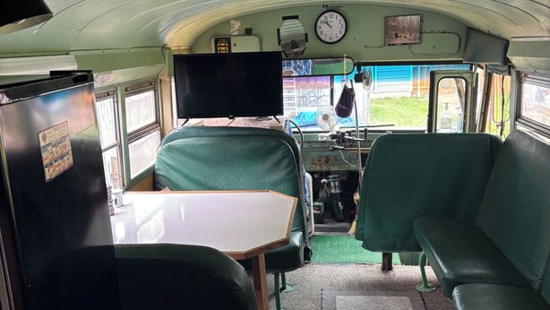 Interior compacto de um veículo recreativo, com uma mesa de jantar, dois sofás verdes, uma pequena cozinha e um relógio na parede.