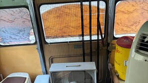 Espaço interior compacto com vaso sanitário, pequeno ventilador, aquecedor de água e janela com vista para paisagem desértica.