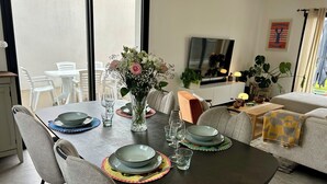 A dining area with a table set for four, chairs, a vase with flowers, and a TV in the background. - Ti Pépin - Contemporary house - 5 min from the beach - Rated 3* furnished for tourism (Fouesnant)