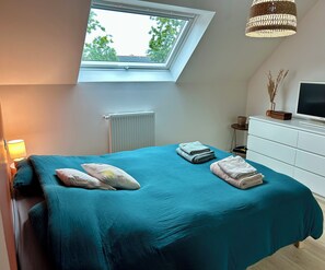 Ein Schlafzimmer mit Oberlicht, einem Bett mit türkisblau gestreuter Bettwäsche, einem Heizkörper, einem Fernseher und einer Kommode.