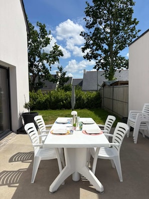 An outdoor patio with a white table set for six, surrounded by white chairs, under a clear sky.