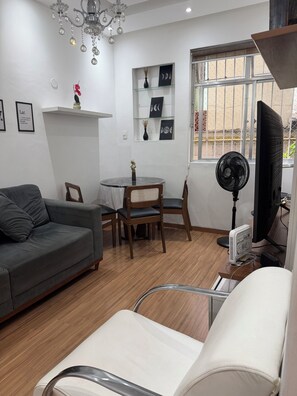 Ein Wohnzimmer mit grauem Sofa, einem Esstisch mit Stühlen, einem Fernseher, einem Ventilator und einem Fenster mit Aussicht.