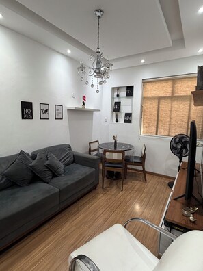 A modern living room with a grey sofa, a dining table with chairs, a chandelier, and a large window with blinds. - Smart & Cozy: Best Location in Botafogo | Steps to Metro & Shops | Sleeps 6 (Rio de Janeiro)