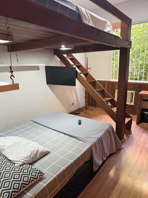Ein Schlafraum mit Etagenbetten, einem Fernseher, einer Holztreppe und einem Fenster mit Blick auf Grünflächen.