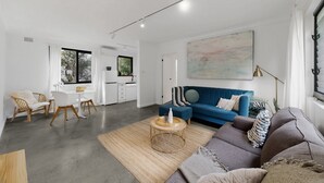 A modern living room with a blue sofa, a round coffee table, and a dining area with a white table and chairs. - Beachside 1-bed Retreat With Parking Cronulla (Sydney)