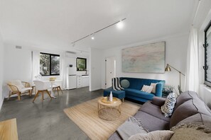 Ein modernes Wohnzimmer mit einem blauen Sofa, einem runden Couchtisch und einem Essbereich mit weißem Tisch und Stühlen.