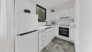 A modern kitchen with white cabinetry, a dishwasher, a stove, and a window with blinds. - Beachside 1-bed Retreat With Parking Cronulla (Sydney)