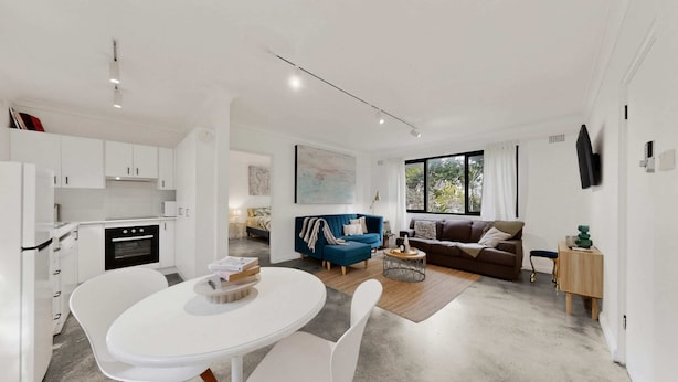 A modern kitchen with white cabinets, a dining table with chairs, a living area with a sofa and a coffee table, and a TV mounted on the wall. - Beachside 1-bed Retreat With Parking Cronulla (Sydney)