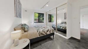 A modern bedroom with a large bed, bedside tables, a mirror, and a view of the outdoors. - Beachside 1-bed Retreat With Parking Cronulla (Sydney)