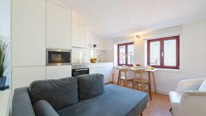Ett kompakt bostadsutrymme med ett litet kök, en soffa, ett matbord och ett fönster med gardiner.