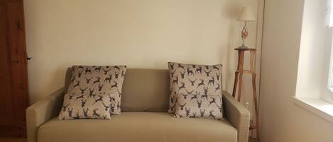 Un canapé beige agrémenté de coussins à motifs, un plancher en bois, une lampe et une fenêtre avec des rideaux.