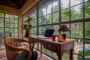 Ein Holztisch mit einem Laptop, einer roten Tasse und einer Lampe, nahe großen Fenstern mit Blick auf üppiges Grün.