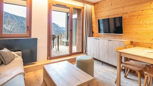 A modern room with a sofa, a wooden table, a TV mounted on the wall, and a sliding glass door leading to an outdoor area with a view of the mountains. - Rental Near Vaujany Ski Lifts (Vaujany)