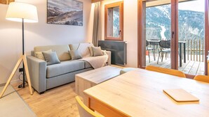A cozy living area with a sofa, a coffee table, a lamp, a TV, and a view of a mountainous landscape through large windows. - Rental Near Vaujany Ski Lifts (Vaujany)
