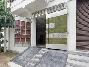 A building entrance with a ramp leading to a door, a closed shutter on the right, and a tree to the left. - Classic With Balcony Chhatikara Char Dham (Mathura)