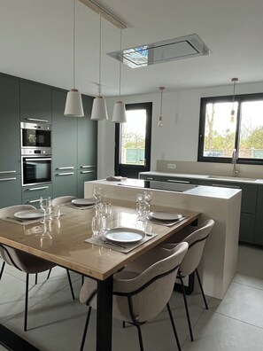 A modern kitchen with a central island, built-in appliances, pendant lighting, and a view through a window. - Le pressoir, calm and nature - 5min Honfleur (Équemauville)