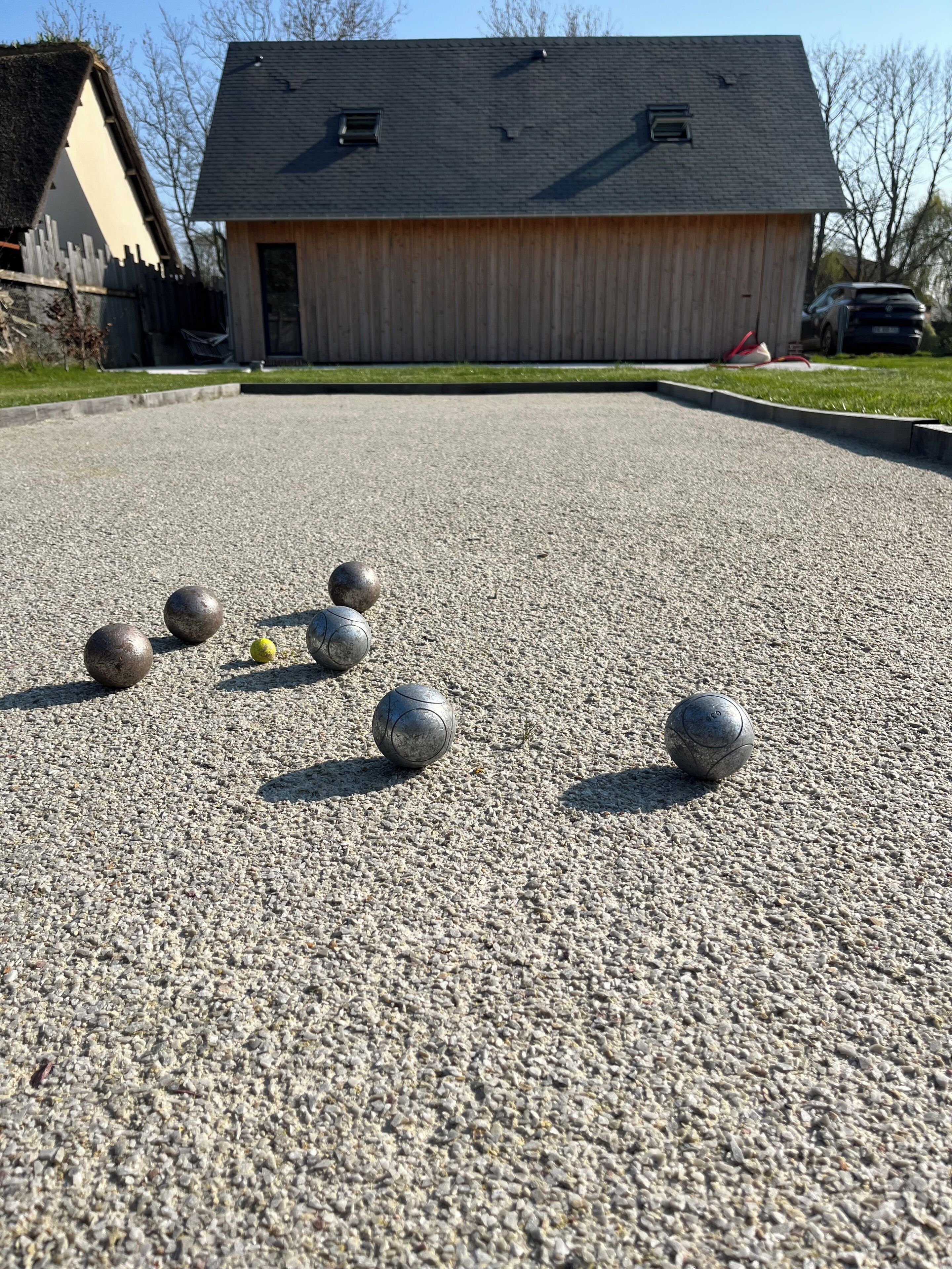 Une cour avec un bâtiment en bois, une surface recouverte de gravier et plusieurs boules métalliques.