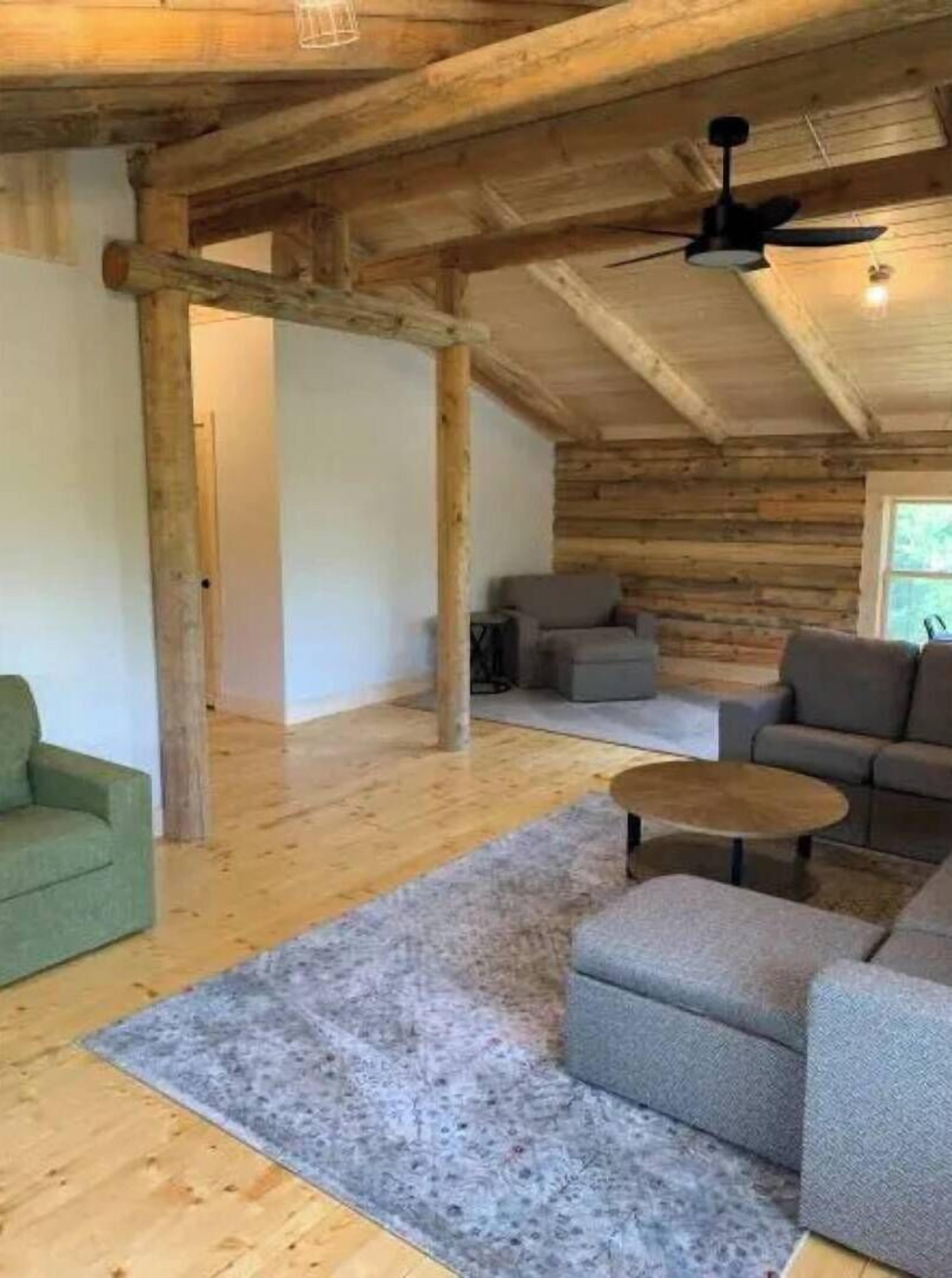 Interior de uma cabana de troncos com teto de madeira, um ventilador de teto, um sofá cinza, uma poltrona verde e uma pequena mesa redonda.