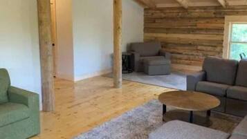 L’intérieur d’une cabane en rondins, avec un plafond en bois, un ventilateur de plafond, un canapé gris, un fauteuil vert et une petite table ronde.