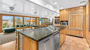 Une cuisine moderne avec des meubles en bois, un grand îlot central, des appareils électroménagers en acier inoxydable et une vue sur l’extérieur.