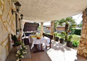 Eine überdachte Terrasse mit Tisch und Bank, Steinmauern, Topfpflanzen und einem Grill.