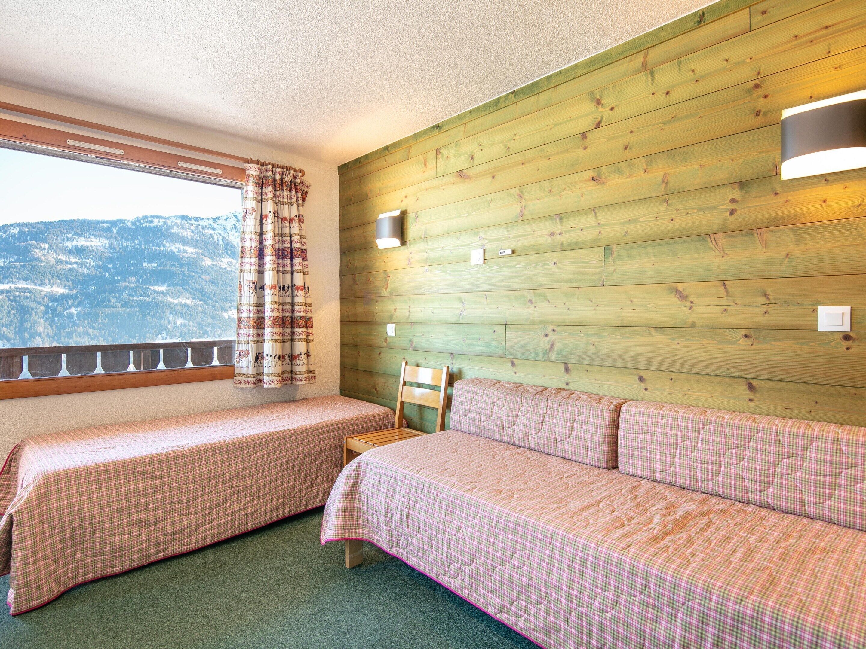A room with two beds, a wooden wall, a window with a view of mountains, and a balcony.